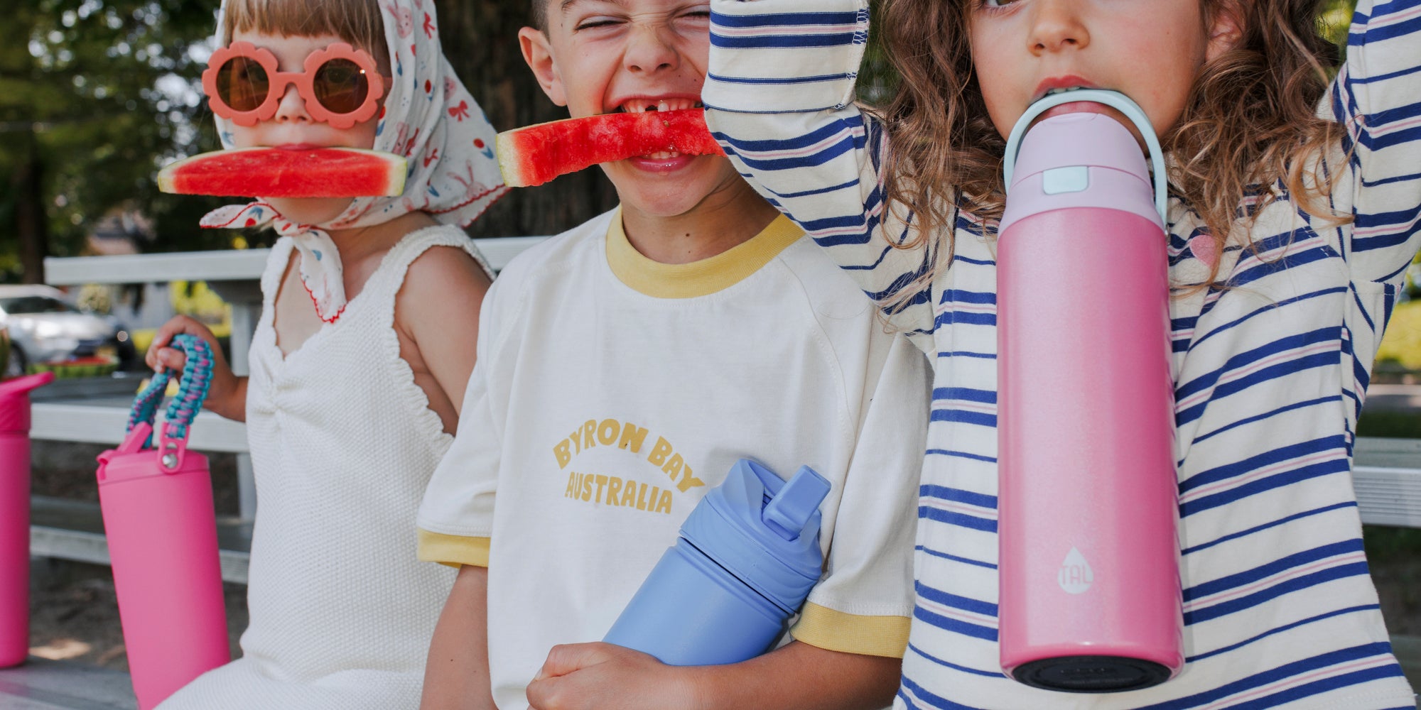 Children sitting on a bench with colorful water bottles and toys outdoors.